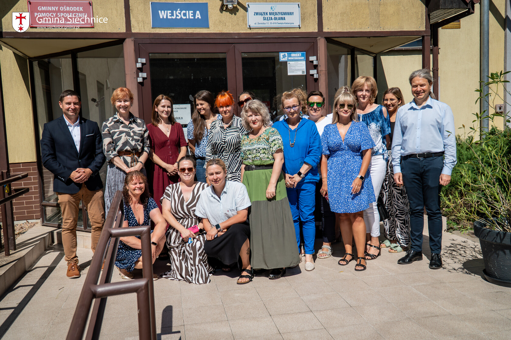 Grupowe zdjęcie pracowników i przedstawicieli gminy przed wejściem do budynku, gdzie działało GOPS, a obecnie mieści się Centrum Usług Społecznych w Siechnicach. Osoby pozują do zdjęcia, uśmiechają się i stoją na tle drzwi oznaczonych szyldem „WEJŚCIE A” oraz tablicami informacyjnymi.