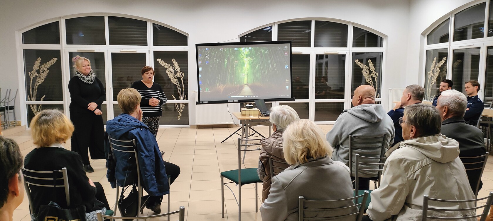 Spotkanie dla seniorów w dużej sali. Przed grupą siedzących osób przemiawia policjant. 