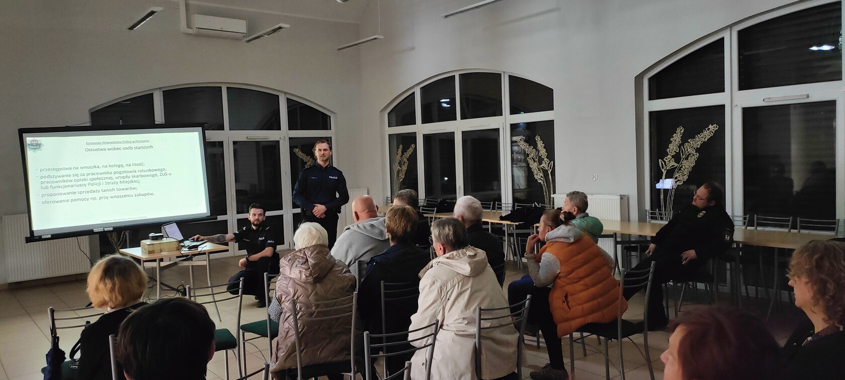 Spotkanie informacyjno-edukacyjne dla seniorów prowadzone przez policjantów. Uczestnicy słuchają prezentacji o zagrożeniach związanych z oszustwami. Slajd przedstawia sposoby działania oszustów wobec osób starszych.