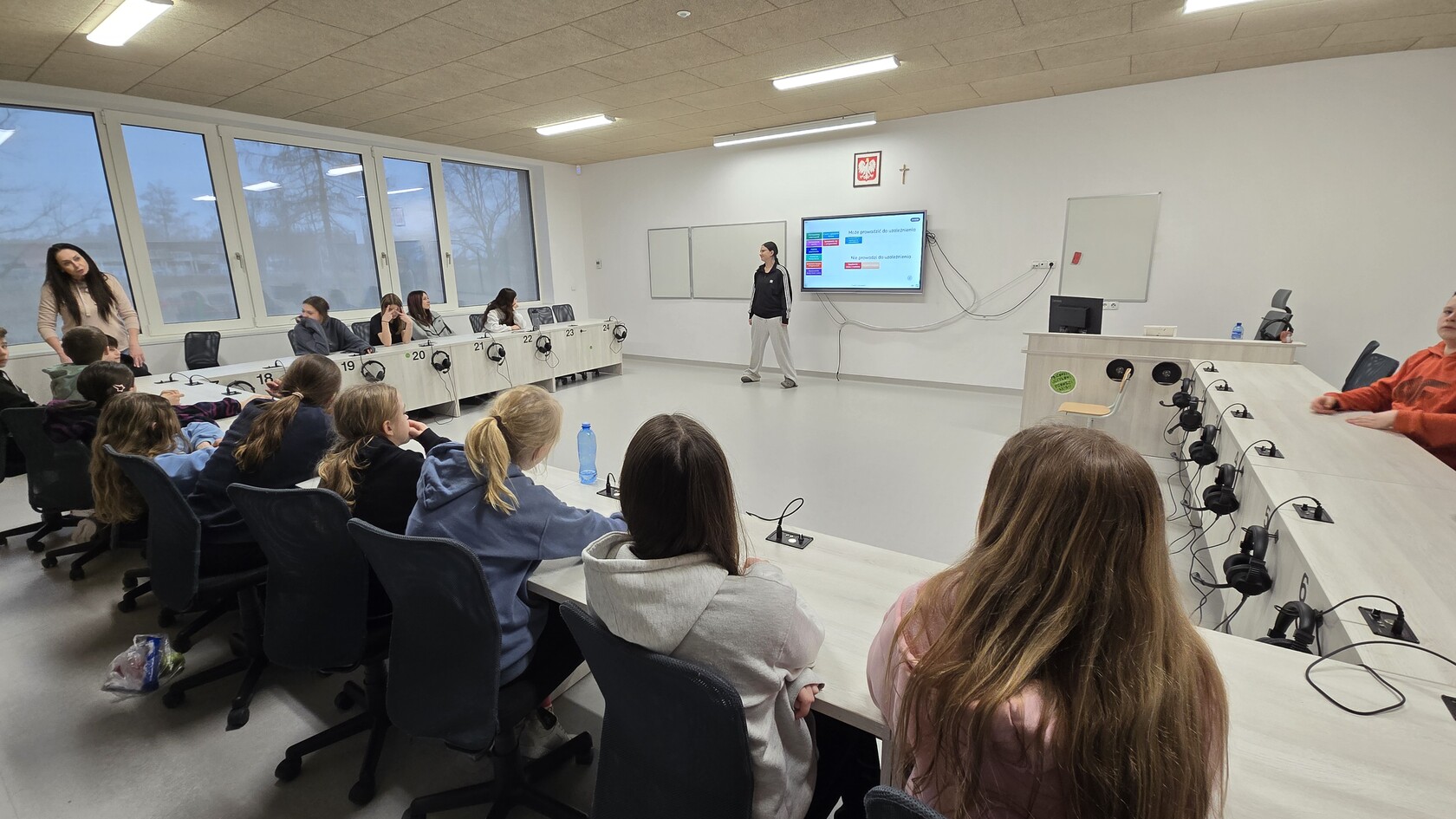 Grupa uczestników półkolonii CUS bierze udział w zajęciach edukacyjnych w sali konferencyjnej. Prowadząca omawia prezentację wyświetlaną na ekranie.